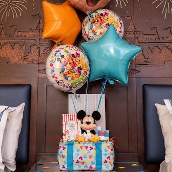 A Mickey's Magical Birthday Celebration decor package displayed on top of a bed features a gift bag overflowing with balloons, a Mickey Mouse plush, a birthday card, a sweet treat, a bag and confetti